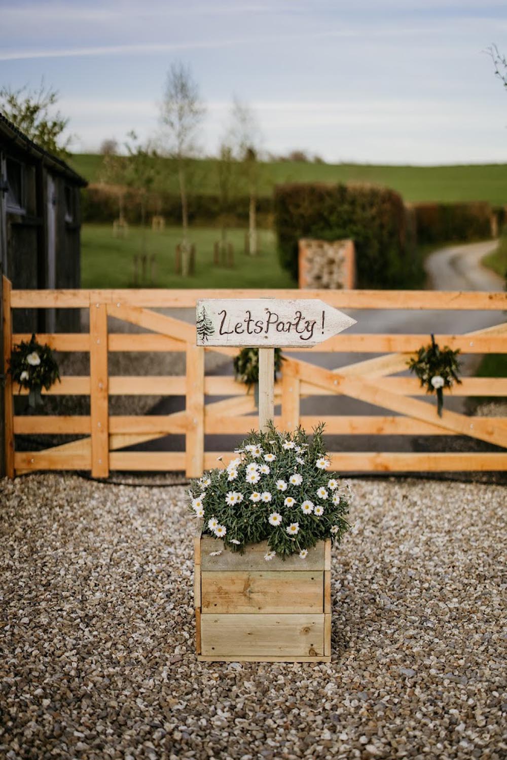Wooden boxes with a "Let's Party!" sign, set before a wooden gate and gravel path.